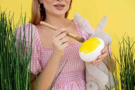 Cropped smiling pregnant woman in light pink dress near high green fresh raw grass, with rabbit toy coloring Easter eggの写真素材