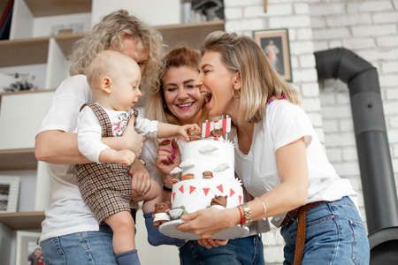 Three smiling beautiful middle-aged and young women congratulate cute little one-year-old boy touching macaroon cake.の写真素材