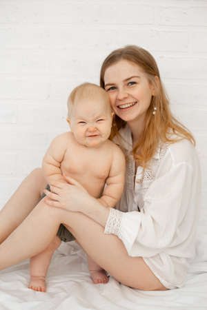 Vertical of young barefoot blond woman with little naked baby in white bedroom, after cleaning procedures. Mother caringの写真素材