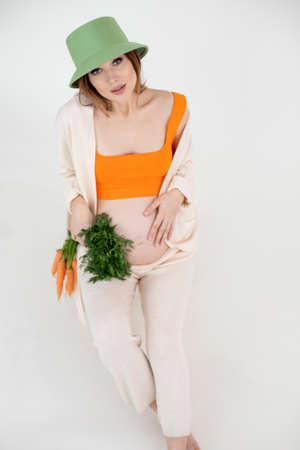 Young gorgeous pregnant woman with long dark hair wear green bucket hat, sitting, holding belly and bunch of carrots.の写真素材