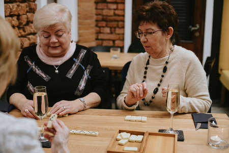 Two smiling and serious senior dark-haired, fair-haired women sit at table with glasses of champagne playing dominoes.の写真素材