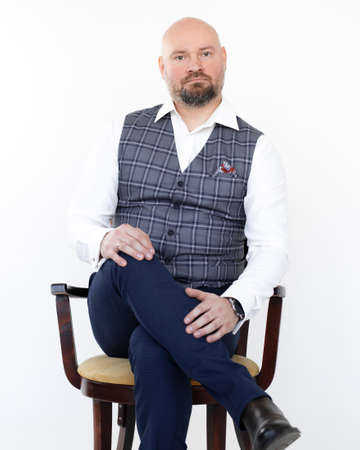 Portrait of elegant middle-aged businessman in grey vest, blue jeans, white shirt sitting on chair...