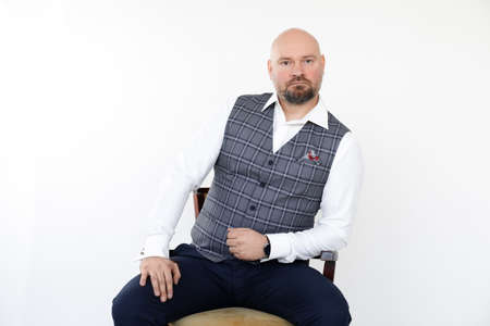 Portrait of bald middle-aged businessman in grey vest, blue jeans, white shirt sitting, leaning elbow on wooden chair.の写真素材