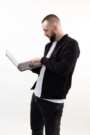 Young engaged bearded man with short dark hair wearing black jacket, jeans and white T-shirt, standing, using laptop.の写真素材