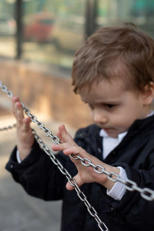 Vertical photo of cute little boy in black jacket standing on the street, holding and touching metal...
