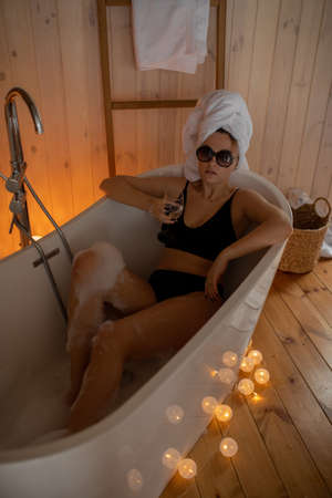 Portrait of woman in black underwear, sunglasses and white towel on head spending weekend at home. Taking bath with glass of wine in cozy bathroom with garland. Home rest and bodycare conceptの写真素材