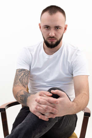Young confident bearded man with short dark hair in white T-shirt, black jeans, sitting on wooden...