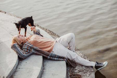Young blonde woman in warm clothes lying on river bank with pretty puppy on cool sunny day, relaxing and enjoying side view. Outdoors weekend activity, love and care for animals, water backgroundの写真素材