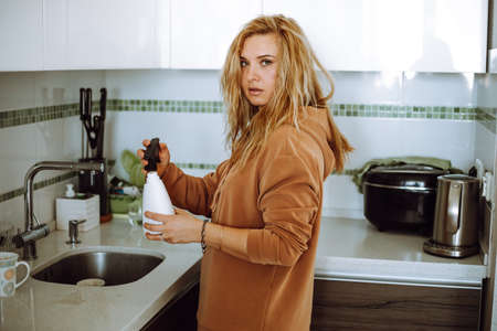 Blonde young woman in home clothes hold and fill flower sprayer with tap water in modern kitchen...