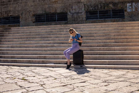 Blond woman in sunglasses sit on luggage on big stairs near stone building. Searching for electronic tickets and mapsの写真素材
