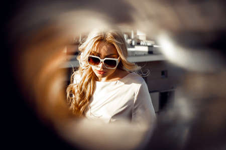 Portrait of young woman with long wavy fair hair wearing beige silk blouse, sunglasses, sitting on...