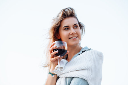 Portrait of happy woman holding in hand wine glass and drinking red wine on white background closeup, walking along embankment in resort city at sunset. Sea voyage, boat mooring, relaxing leisureの写真素材
