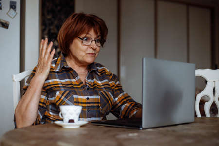 Emotional mature lady in glasses looking at laptop screen at table in room. Advanced pensioner...