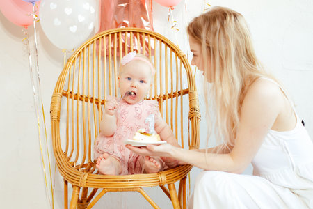 Portrait closeup of young mother and baby girl sitting and eating cake in holiday dress on white...