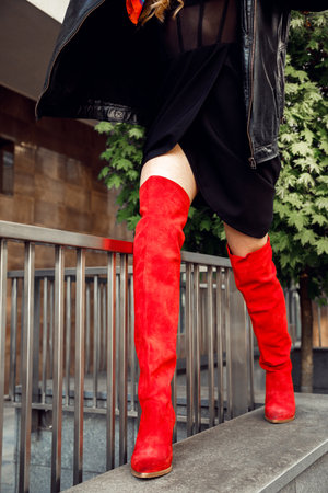 Cropped photo of slim woman wearing short black dress, leather jacket, red high boots walking near building in city.の写真素材