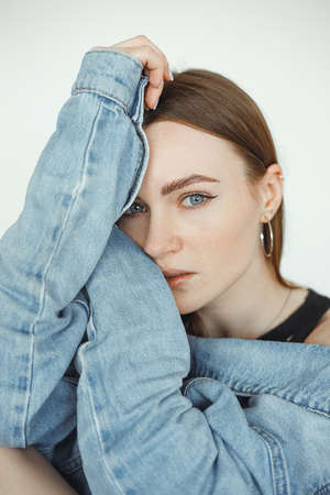 Vertical of calm, serious dark-haired woman looking at camera, wearing denim jeans jacket. Hugging...