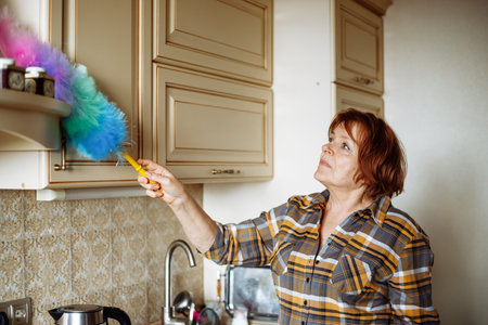Portrait of senior attentive woman standing in beige kitchen, wiping dust off cupboards with soft colorful duster.の写真素材