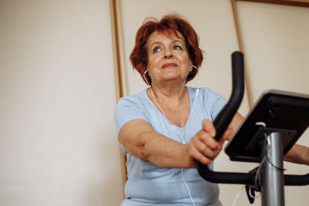 Portrait of elderly woman with dark hair, make-up wearing blue T-shirt, sitting on stationary bicycle, doing exercises.の写真素材