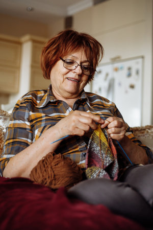 Portrait of senior woman with dark hair wearing checkered shirt, glasses, sitting on sofa, knitting...
