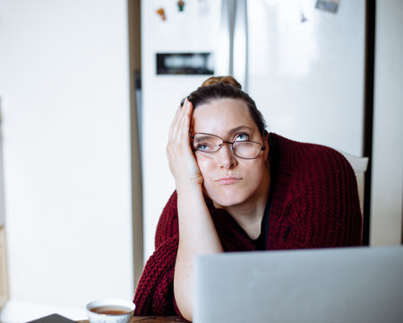 Portrait of middle-aged dissatisfied woman with dark hair in bun wearing cardigan, glasses sliding up, leaning on table.の写真素材