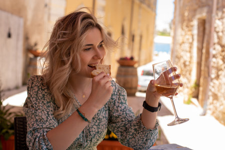 Beautiful blond woman sitting at outdoor cafe in hot summer day. Eating bread, drink white wine. Sea...