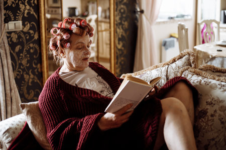 Side view of elderly woman applying pink hair-curlers, facial mask, wearing vinous cardigan, sitting, reading book.の写真素材