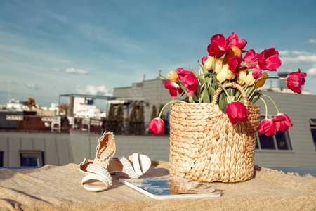 Composition of straw bag with bunch of pink, yellow flowers tulips, beige sandals and magazine on linen cloth on roof.の写真素材