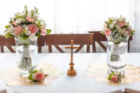 Soft bridal bouquets of fresh flowers in glass vases and church cross on celebratory table in restaurant. Elegant attribute and tradition. Beautiful and romantic wedding, luxurious celebrationの写真素材