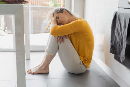 Depressed 30 years woman sitting on kitchen floor with hugging knees. Loneliness, sadness. Mental health, drug abuse and social issue concept.の写真素材