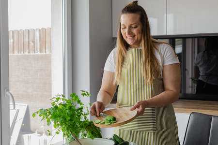 Smiling woman 40 years old in apron cutting cucumbers on cutting board in kitchen at home. Woman cookingの写真素材