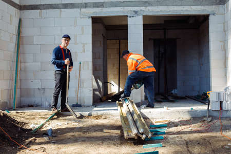 Mature workers, men in protective coveralls using chainsaw cutting wood bars and steel tubes, girder near building wallの写真素材