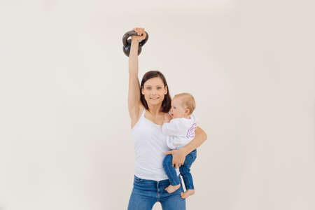 Young athletic smiling woman lifting up kettlebell and holding little baby, white background. Strength workout and weightlifting, pumping up muscles, mother and child. Sport and healthy lifestyleの写真素材