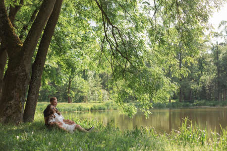 Young couple falling in love sitting on grass under tree near lake in park, looking on each other. Young man and womanの写真素材