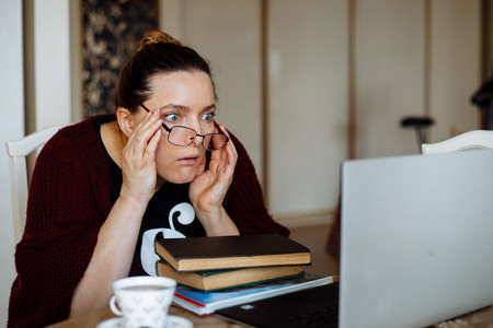 Surprised middle-aged woman sitting at table near books, looking at laptop, adjusting glasses, not...