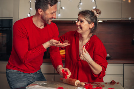 Young cute woman and man dressed in red clothes laugh merrily while baking cookies. Christmas moments, happy young couple. Home cozy kitchen.の写真素材
