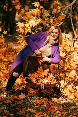 Stylish blonde inknitted sweater and bright lilac coat smiles and shows pile of fallen orange autumn...