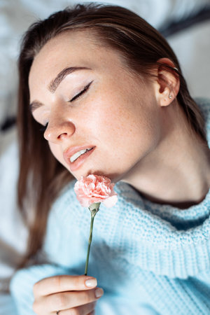 Sensual young woman with beautiful smile in blue sweater lying on bed with flower in hand. Portrait of dreamy lady restingの写真素材