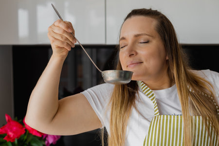 Closeup plump blonde woman in apron cooking and smelling tasty soup in ladle with close eyes....