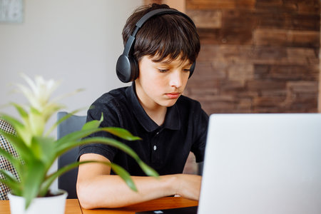 Portrait of concentrated teenager boy wearing black T-shirt, headphones sitting at desk, using...