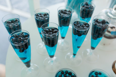 Serving row of glasses of refreshing colorful drinks with blueberry closeup, white background. Pouring sparkling wine into glasses, event celebration. Banquet hall, festive party, luxurious serviceの写真素材