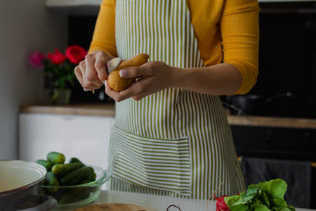 Cropped woman in apron peeling potato and chop fresh vegetables. Cooking mixed summer salad or soup. Healthy eating foodの写真素材