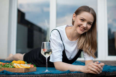 Happy young woman lying on blue plaid on terrace with glass of white wine and snacks closeup....