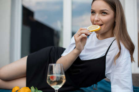 Happy young woman lying on blue plaid on terrace with glass of white wine and eating orange closeup. Drinking alcohol drink and enjoying outdoor recreation. Home party, vacation and loungeの写真素材
