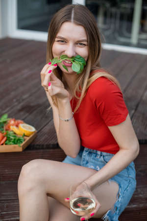 Cheerful young woman sitting on terrace with glass of white wine and plate of fresh greens and snacks closeup. Drinking alcohol drink and enjoying outdoor recreation. Home party, vacation and loungeの写真素材