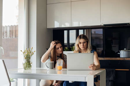 Two women sitting at table in light modern kitchen, working together on laptop and drinking fresh...