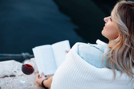 beautiful young woman is sitting on pier with glass of red wine and book having great time on vacation , happy evening.の写真素材