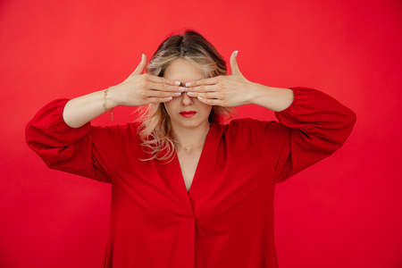 Portrait of hiding woman in red outfit with perfect bright makeup closing eyes by hands isolated on red background. Avoiding problems, fear, giving under stress. Fashion and beauty industryの写真素材