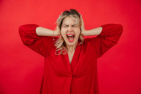 Portrait of loudly screaming woman in red outfit with perfect bright makeup holding on head by hands isolated on red background. Losing control, giving under stress. Fashion and beauty industryの写真素材