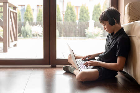 Side view of teenager boy wearing black headphones, sitting on floor, putting laptop on legs,...