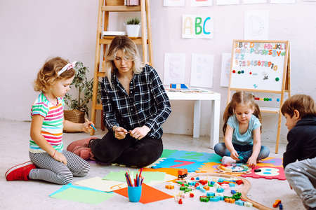 Little kids and educator folding lego and drawing with colored pencils and markers sitting on floor in playroom. Interesting lesson for kindergartners developing creation and intelligenceの写真素材